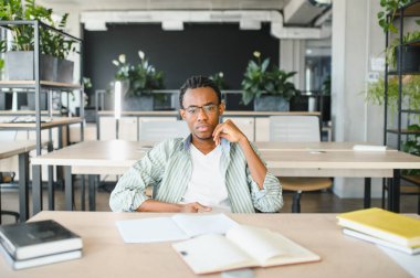 Kitaplarla çevrili ve gelecek sınavlara hazırlanan parlak, modern bir üniversite kütüphanesinde derslerine konsantre olan genç adam.
