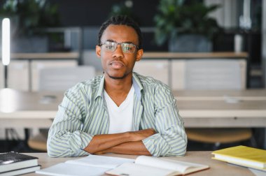 Odaklanmış Afro-Amerikan üniversite öğrencisinin portresi açık modern kütüphanede ya da sınıfta çapraz kollu, kitaplar ve defterlerle çevrili bir şekilde masada oturuyor.