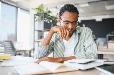 Genç Afrikalı Amerikalı erkek öğrenci kitap okuyor ve not alıyor, üniversite kütüphanesinde sınavlara hazırlanıyor.