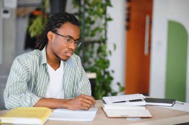 Genç Afrikalı Amerikalı erkek öğrenci üniversite kütüphanesinde okurken bir not defterine notlar yazıyor.