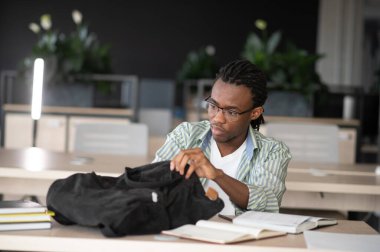 Modern kütüphanede kitap ve defterlerle masa başında otururken sırt çantasında araştırma yapan Afro-Amerikan üniversite öğrencisi.