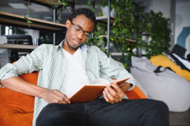 Çağdaş bir kütüphane ortamında kitap ve bitkilerle çevrili ders kitabı okuyan, derslerine konsantre olan genç siyahi bir adam.