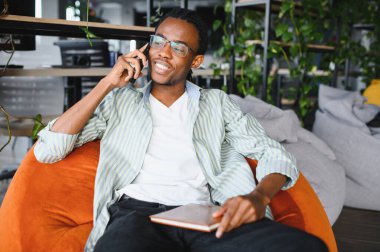 Gülümseyen Afro-Amerikan öğrenci akıllı telefondan konuşuyor, elinde bir defter, üniversite kütüphanesinde turuncu puf koltukta oturuyor.