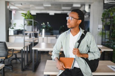 Düşünceli Afrikalı Amerikalı öğrenci, elinde kitaplarla modern üniversite kütüphanesinde dikilirken gözlerini kaçırıyor.