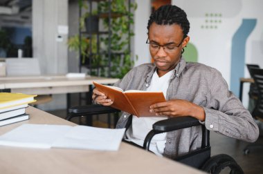 Tekerlekli sandalye kullanan genç bir Afrikalı Amerikalı parlak, modern bir ofiste çalışırken kitap okuyor.