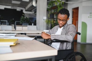 Tekerlekli sandalyedeki genç siyahi adam, kitap okuyor ve parlak, modern bir ofiste eğitim ve kişisel gelişime odaklanıyor.