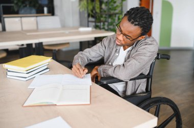 Tekerlekli sandalyedeki Afrikalı Amerikalı öğrenci, modern çalışma ortamındaki kitaplarla çevrili bir masada notlar yazmaya odaklanıyor.