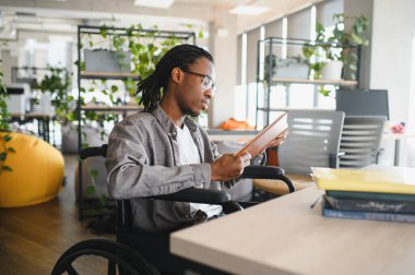 Genç Afro-Amerikalı bir adam tekerlekli sandalye kullanıyor. Modern, erişilebilir bir ofis ortamında tablet okuyor. Kapsama alanı ve odaklanmış çalışma gösteriyor.