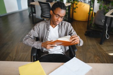 Tekerlekli sandalyedeki Afrikalı Amerikalı öğrenci masa başında kitap okuyor, modern bir sınıf ortamında çalışmalara odaklanıyor.