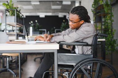 Tekerlekli sandalyedeki genç siyahi profesyonel dizüstü bilgisayarda çalışıyor ve not alıyor, iş yerinde kapsayıcılığı ve erişilebilirliği gösteriyor