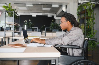 Çağdaş bir ofiste dizüstü bilgisayarla çalışan, bitkilerle ve kitaplarla çevrili, tekerlekli sandalyedeki Afrikalı Amerikalı öğrenci,