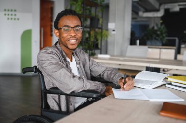 Tekerlekli sandalye kullanan mutlu genç siyahi adam, modern bir kütüphanede çalışırken not alıyor.