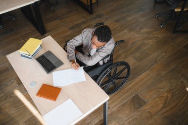 Tekerlekli sandalye kullanan Afrikalı Amerikalı bir öğrenci, bir masada dizüstü bilgisayar ve kitaplarla notlar yazıyor, eğitim ve erişilebilirliğe odaklanıyor.
