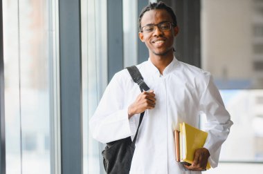 Laboratuvar önlüğü giyen genç siyah bir adam, kitap ve sırt çantası taşıyor, parlak bir üniversite koridorunda yürürken güvenle gülümsüyor.