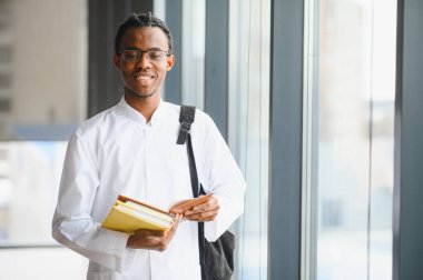 Üniversite koridorunda kitap ve sırt çantası tutarken gülümseyen Afro-Amerikan tıp öğrencisi.