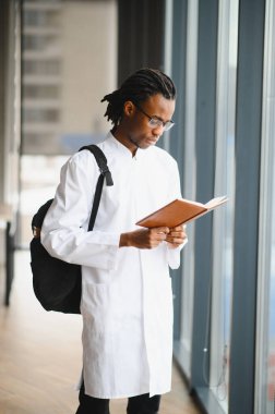 Odaklanmış Afro-Amerikan tıp öğrencisi ders kitabı okuyor, parlak bir üniversite koridorunda sınavlara hazırlanıyor.