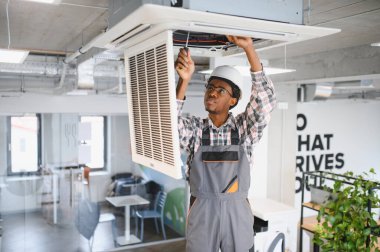 Genç Afro-Amerikan erkek teknisyen güvenlik kaskı ve tulum giyiyor, tavan monte edilmiş klima ünitesini tamir etmek için tornavida kullanıyor.