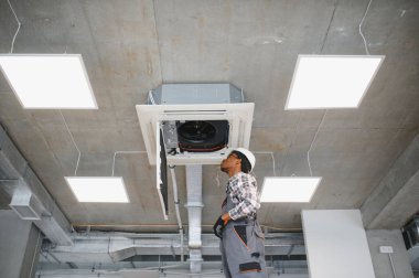 Güvenlik teçhizatı kullanan Afrikalı Amerikalı teknisyen modern endüstriyel alanda bir tavan havalandırma ünitesini tamir etmeye çalışıyor.