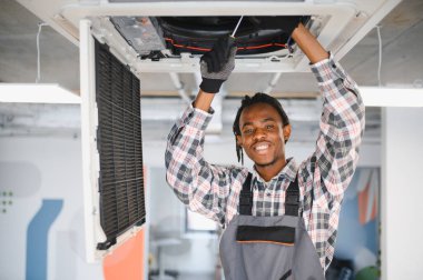 Genç Afrikalı Amerikalı işçi havalandırma sistemini tamir ederken tornavida kullanıyor ve işte gülümsüyor.