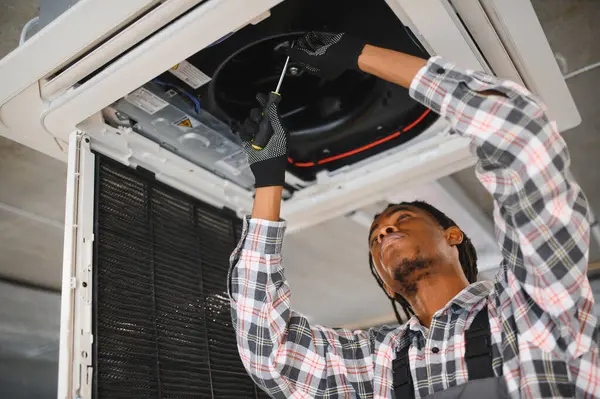 Genç Afro-Amerikan bakım işçisi bir tornavidayla havalandırma ünitesini tamir ediyor, kapalı alanda verimli ve yetenekli servislere odaklanıyor.