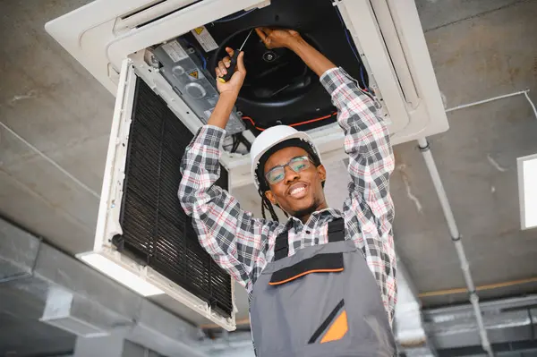 Genç Afro-Amerikan bakım işçisi bir tornavidayla havalandırma sistemini tamir ediyor, içeride ideal soğutma ve konfor sağlıyor.