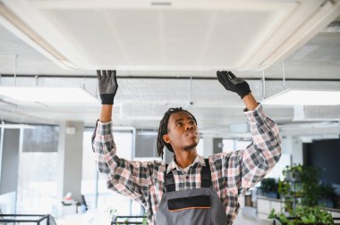 Genç Afrikalı Amerikalı işçi ofis binasında havalandırma sistemini tamir ediyor.