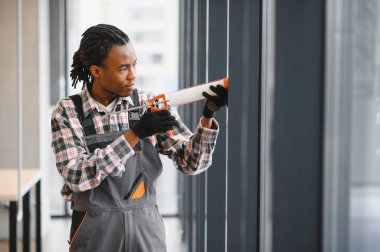 Afrika kökenli Amerikalı işçiler modern ofis alanındaki pencere kurulumunda, hava geçirmez mühürleme sağlanırken, bir kabartma tabancasıyla dolgu macunu uyguluyorlar.