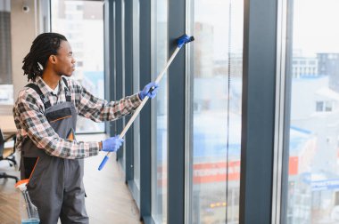 Bir ofis binasının camlarını temizleyen genç adam temiz ve hijyenik bir ortam sağlamak için cam sileceği ve temizlik sıvısı kullanıyor.