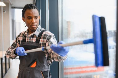 Geniş ofis pencerelerini silmek için cam sileceği kullanan üniformalı bir işçi, bakım ve temizlikte uzmanlığı vurguluyor
