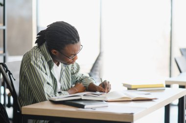 Ders kitaplarıyla çevrili, parlak bir kütüphane ortamında sınava hazırlanırken, bir not defterine notlar yazan odaklanmış erkek üniversite öğrencisi