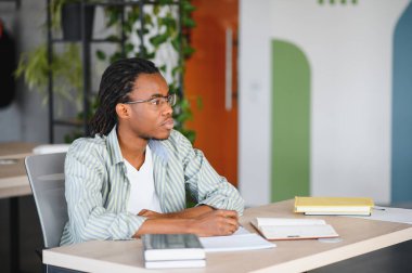 Genç Afrikalı Amerikalı öğrenci notlar alıyor ve modern bir üniversite kütüphanesinde ya da ortak çalışma alanında okuyor.