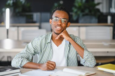 Genç Afrikalı Amerikalı öğrenci üniversite kütüphanesinde okurken gülümsüyor ve notlar alıyor.