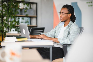 Çalışmalarına konsantre olan genç bir adam, parlak, modern bir üniversite kütüphanesinde dizüstü bilgisayarla yazı yazıyor.