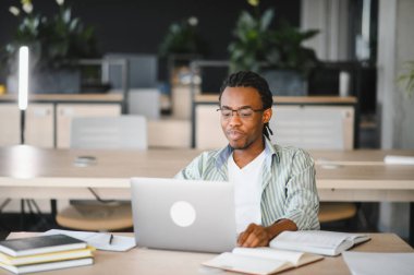 Genç Afrikalı Amerikalı erkek öğrenci dizüstü bilgisayar kullanıyor ve kitap okuyor. Üniversite kütüphanesinde sınava hazırlanıyor.