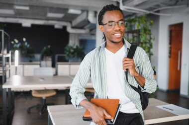 Kitap ve sırt çantası taşıyan mutlu öğrenci, parlak modern kütüphanede üniversite hayatının ve akademik uğraşların tadını çıkarıyor.