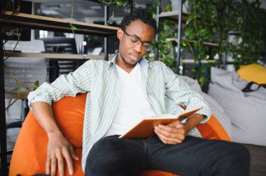 Genç Afrikalı Amerikalı erkek öğrenci üniversite kütüphanesinde puf minderinde otururken kitap okuyor. Sınavlara ve mezuniyete hazırlanıyor.