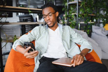 Genç Afrikalı Amerikalı öğrenci üniversite kütüphanesinde puf minderinde oturup sınavlara hazırlanırken akıllı telefon kullanıyor ve kitap tutuyor.