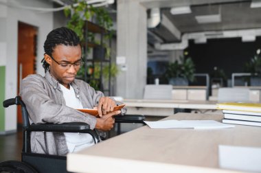 Tekerlekli sandalye kullanan, modern ofiste notlar okuyan, eğitim ve kariyerine odaklanmış genç siyahi bir adam.