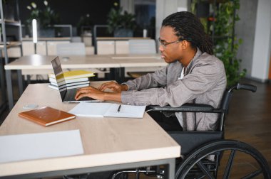 Özürlü genç bir adam, modern bir ofiste dizüstü bilgisayarıyla çalışıyor. İşyerine dahil edilme ve erişilebilirliğini gösteriyor.