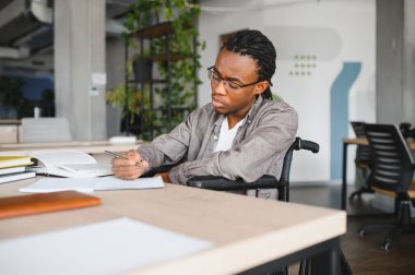 Tekerlekli sandalye kullanan genç siyah bir adam, notlar alıyor ve parlak bir çalışma alanında öğrenim görüyor, eğitimi ve fırsatı kucaklıyor.