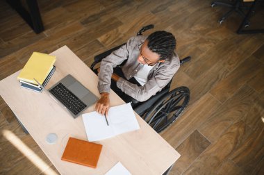 Tekerlekli sandalyedeki genç Afro-Amerikan öğrencinin yüksek açılı görüşü, masada dizüstü bilgisayar kullanırken not alması, eğitime olan bağlılığını göstermesi ve fiziksel sınırlamaların üstesinden gelmesi.
