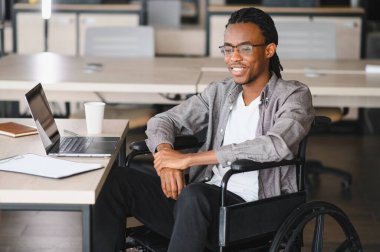 Tekerlekli sandalyede oturan Afrikalı Amerikalı bir öğrenci dizüstü bilgisayarla masa başında çalışıyor, eğitim ortamında kararlılık ve kapsayıcılık sergiliyor