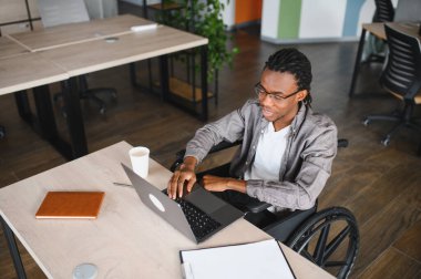 Özürlü genç bir zenci modern bir ofiste çalışıyor, işleri yönetmek ve çevrimiçi bağlantı kurmak için dizüstü bilgisayar kullanıyor.