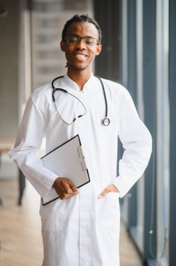 Laboratuvar önlüğü ve steteskop giyen kendine güvenen doktor, elinde sağlık raporu ve hastane koridorunda gülümsüyor.