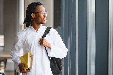 Üniversite koridorunda kitap ve sırt çantası taşıyan kendinden emin Afro-Amerikan tıp öğrencisi pencereden dışarı bakıyor.