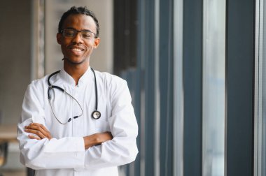 Kollarını kavuşturup gülümseyen, modern bir hastane ortamında beyaz önlük ve steteskop giyen, kendinden emin, genç, Afro-Amerikan erkek bir doktorun portresi.