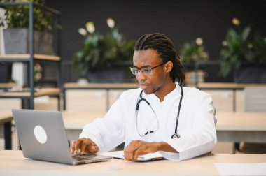 Steteskoplu genç bir erkek doktor, yeşil ve modern dekorlarla çevrili çağdaş bir ofis alanında özenle bir dizüstü bilgisayar üzerinde çalışıyor.