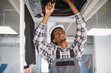 Sert şapkalı ve tulumlu Afrikalı Amerikalı teknisyen tavana monte edilmiş havalandırma ünitesi üzerinde çalışıyor.
