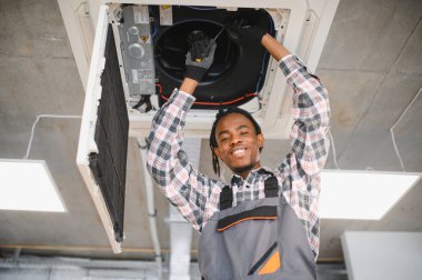 Gülümseyen Afro-Amerikan HVK teknisyeni tornavida kullanarak havalandırma sistemini tamir ediyor.