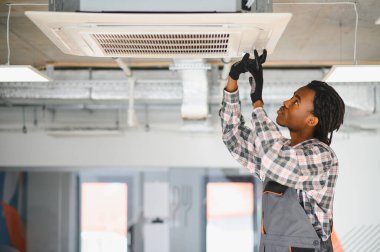 Genç Afro-Amerikan HVK teknisyeni bakım sırasında tavana monte edilmiş kaset tipi klimayı inceliyor.